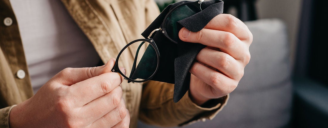 A close-up view shows an individual carefully cleaning a pair of eyeglasses using a black microfiber cloth. The person is wearing a tan jacket and is seated indoors, focusing on the task. The image highlights the hands, glasses, and cleaning cloth, with a neutral and soft color palette. No visible text or numbers are present.