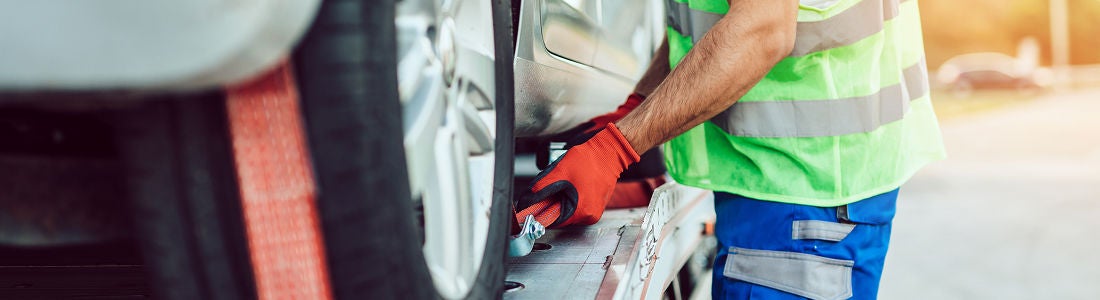 A person in a high-visibility vest and red gloves is securing a car onto a flatbed tow truck. The scene takes place outdoors in daylight, with the worker focused on attaching the vehicle. The image highlights the safety gear and the process of vehicle recovery. No visible text or numbers are present.