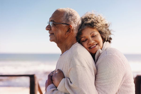 An older couple stands outdoors, warmly embracing each other near the ocean. The woman smiles with her eyes closed, resting her head on the man's shoulder, while he looks contentedly into the distance. Soft daylight and a clear sky create a peaceful, relaxed atmosphere. The scene conveys affection, companionship, and joyful moments in later life.