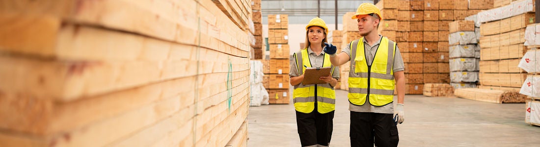 Two workers wearing yellow safety vests and helmets walk through a warehouse filled with large stacks of lumber. One person holds a clipboard and gestures while discussing inventory with a colleague. The setting is industrial, with a focus on organization and teamwork in a lumber storage facility. The mood is professional and collaborative, with neutral and yellow tones dominating the scene.