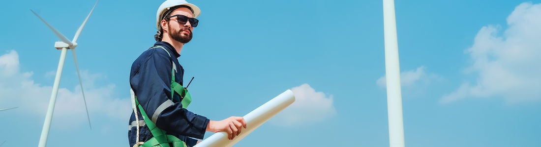 A person wearing a hard hat and safety gear holds a rolled blueprint while standing outdoors near large wind turbines. The setting is a wind farm under a clear blue sky, suggesting renewable energy and engineering activity. The mood is professional and focused, with clean visuals and modern technology elements.