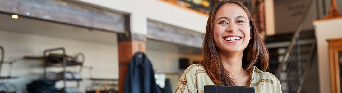 A young adult woman stands indoors holding a tablet device, set against the backdrop of a contemporary retail store. The environment features industrial design elements, visible clothing racks, and a staircase. The overall mood is professional and welcoming, with natural lighting and neutral tones. No visible text or numbers are present in the image.