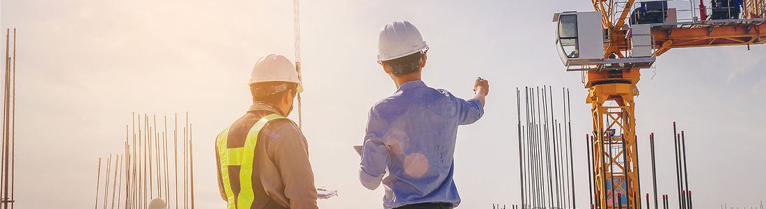 Two adult men wearing safety helmets and vests are seen at a construction site, engaged in conversation. The setting features steel rods, a crane, and bright sunlight, creating a professional and industrious atmosphere. The image captures teamwork and planning in an urban development environment. No visible text or numeric values are present.
