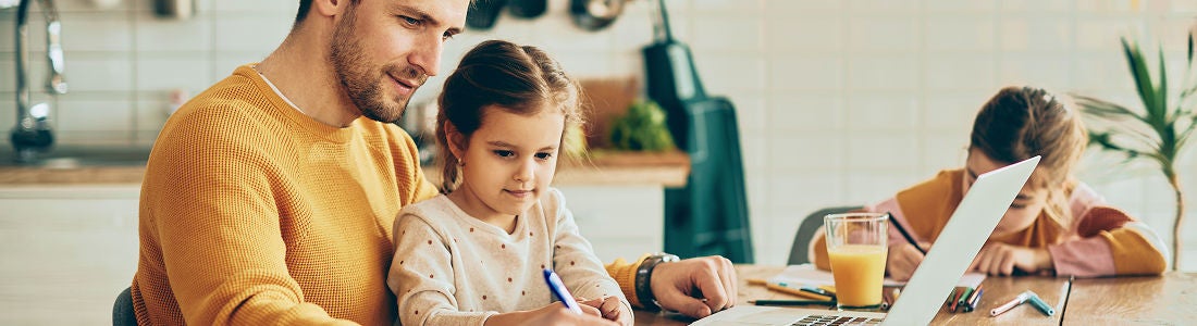 An adult male and two young girls are seated at a kitchen table, engaged in work and study activities. The father assists one child with writing while the other focuses on her own task. A laptop and a glass of orange juice are visible, suggesting a home learning or remote work environment. The setting is bright and casual, with warm tones and a cozy atmosphere.