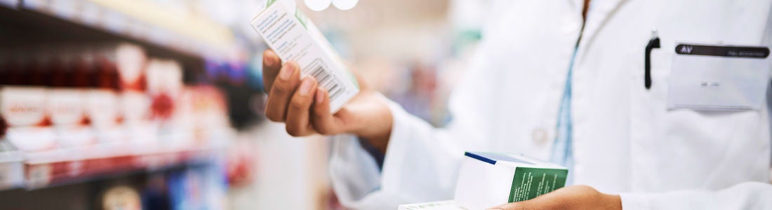 A person in a white lab coat is holding and inspecting two medicine boxes in a pharmacy setting. Shelves filled with various products are visible in the background, suggesting a retail or healthcare environment. The scene conveys a professional and clinical mood, with a focus on healthcare and medication. No visible text or numeric values are clearly identifiable on the boxes.