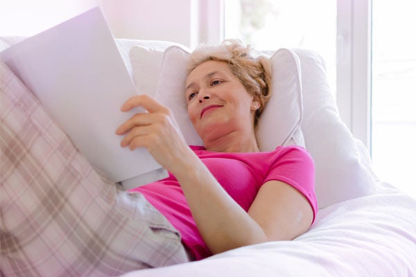 An older woman reclines comfortably on a white sofa while reading a book or magazine. She is wearing a bright pink top and plaid pajama pants, suggesting a cozy, casual moment at home. Soft natural light comes through the window in the background, creating a calm and peaceful atmosphere. The overall scene conveys relaxation, leisure, and quiet personal time.